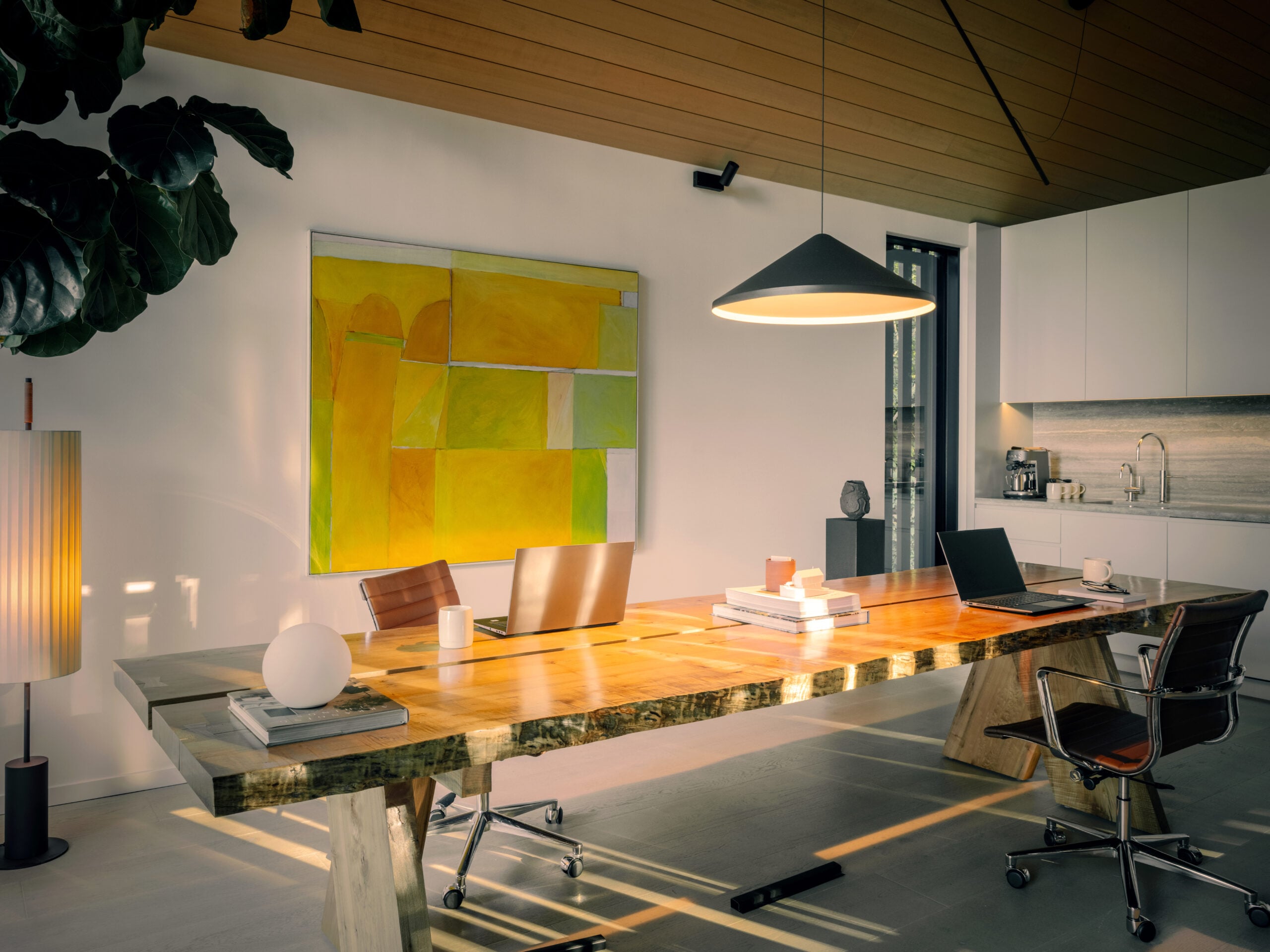 Open workspace with large wood table beneath pendant light with yellow artwork displayed on back wall