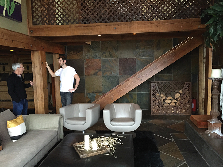 Living room with a stone wall, wooden beams, two grey chairs, a couch, and two men talking.