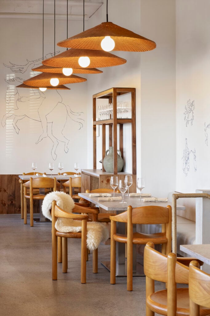 Dining area with light wood pine chairs, sheepskin throws, quartz tables, and a wall mural of a figure on horseback, beneath a row of rattan pendant lights