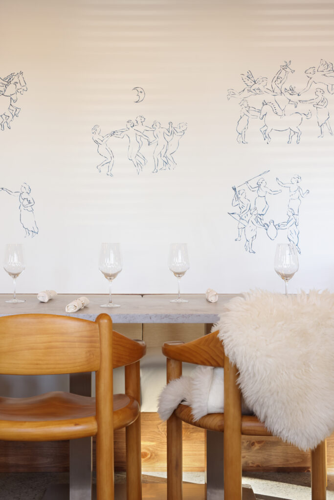 Close-up of a wall mural showing groups of dancing Greek-style figures and a crescent moon, above a dining table with wine glasses and pine chairs