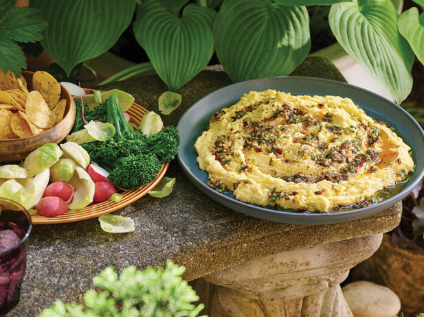 A bowl of creamy corn dip topped with chili oil sits on a stone ledge outdoors, surrounded by lush greenery. Beside the dip, a plate of fresh vegetables and chips is arranged for dipping. The vibrant scene features a relaxed, garden-like atmosphere with a mix of rustic and natural elements.