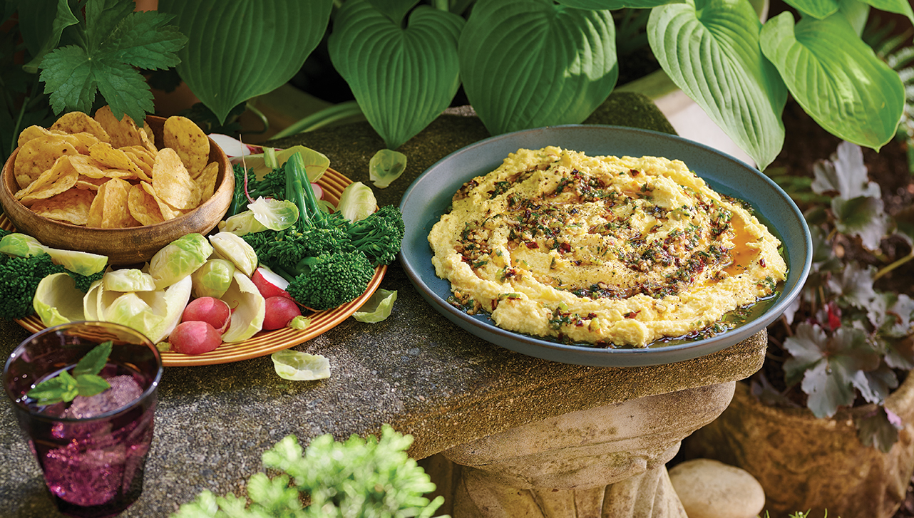 A bowl of creamy corn dip topped with chili oil sits on a stone ledge outdoors, surrounded by lush greenery. Beside the dip, a plate of fresh vegetables and chips is arranged for dipping. The vibrant scene features a relaxed, garden-like atmosphere with a mix of rustic and natural elements.