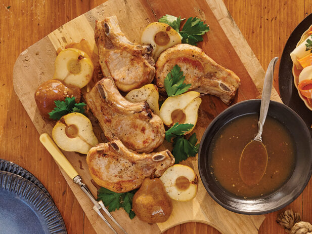 Grilled pork chops are beautifully arranged on a wooden cutting board alongside roasted pears and fresh parsley. A bowl of savoury sauce with a spoon is set nearby, ready for serving. The rustic table setting creates a warm, inviting feel, perfect for a hearty meal.