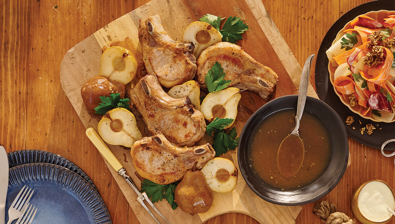 Grilled pork chops are beautifully arranged on a wooden cutting board alongside roasted pears and fresh parsley. A bowl of savoury sauce with a spoon is set nearby, ready for serving. The rustic table setting creates a warm, inviting feel, perfect for a hearty meal.