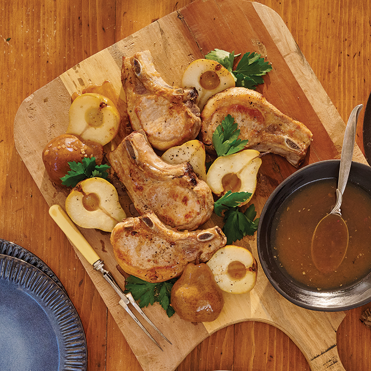 Grilled pork chops are beautifully arranged on a wooden cutting board alongside roasted pears and fresh parsley. A bowl of savoury sauce with a spoon is set nearby, ready for serving. The rustic table setting creates a warm, inviting feel, perfect for a hearty meal.