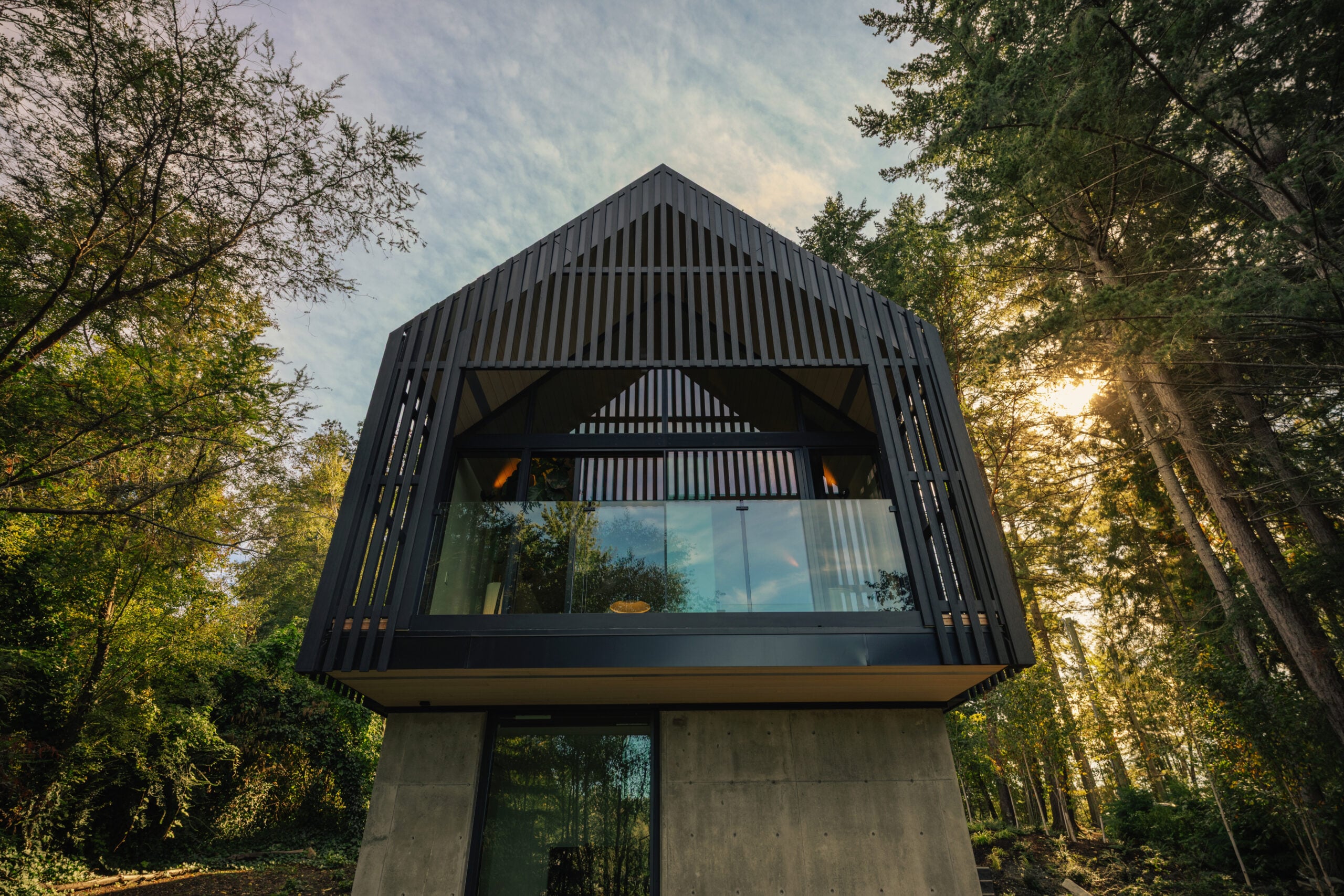 Low-angle rear view of the elevated cedar studio, with a concrete base and deck with a glass guardrail.