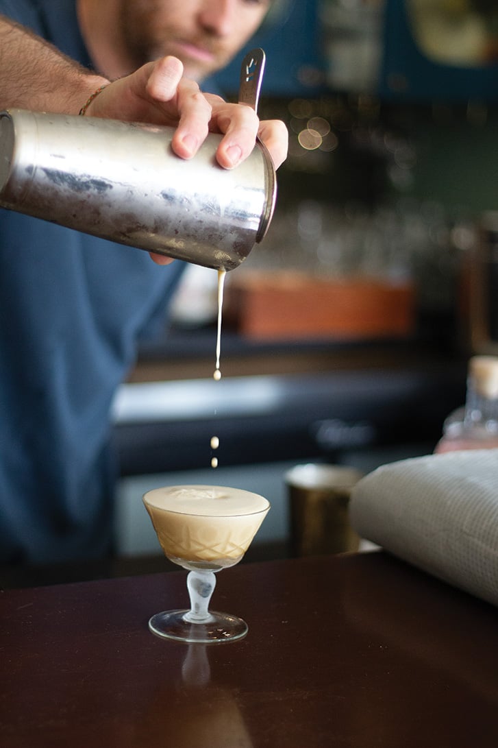 Bartender pouring the Chez Solange cocktail from a metal shaker into a coupe glass.
