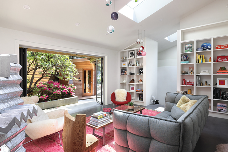 Bright living room with a grey sofa, pink rug, and built-in shelves, opening to a garden with blooming flowers.