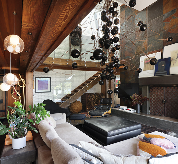 Living area with wooden beams, a stone fireplace, and eclectic hanging lights, featuring a beige sofa and colourful rug.