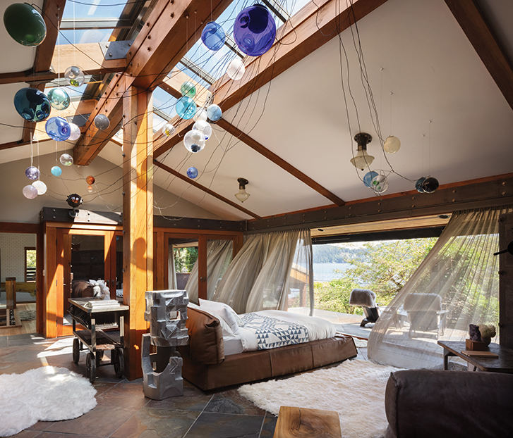 Bedroom with wooden beams and large windows overlooking a lake, featuring a brown bed and colourful hanging lights.