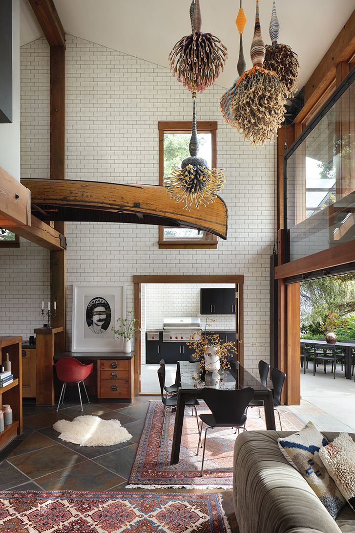 Dining area with wooden beams, colourful rugs, eclectic hanging lights, and a wooden canoe above, opening to a kitchen with white subway tiles.