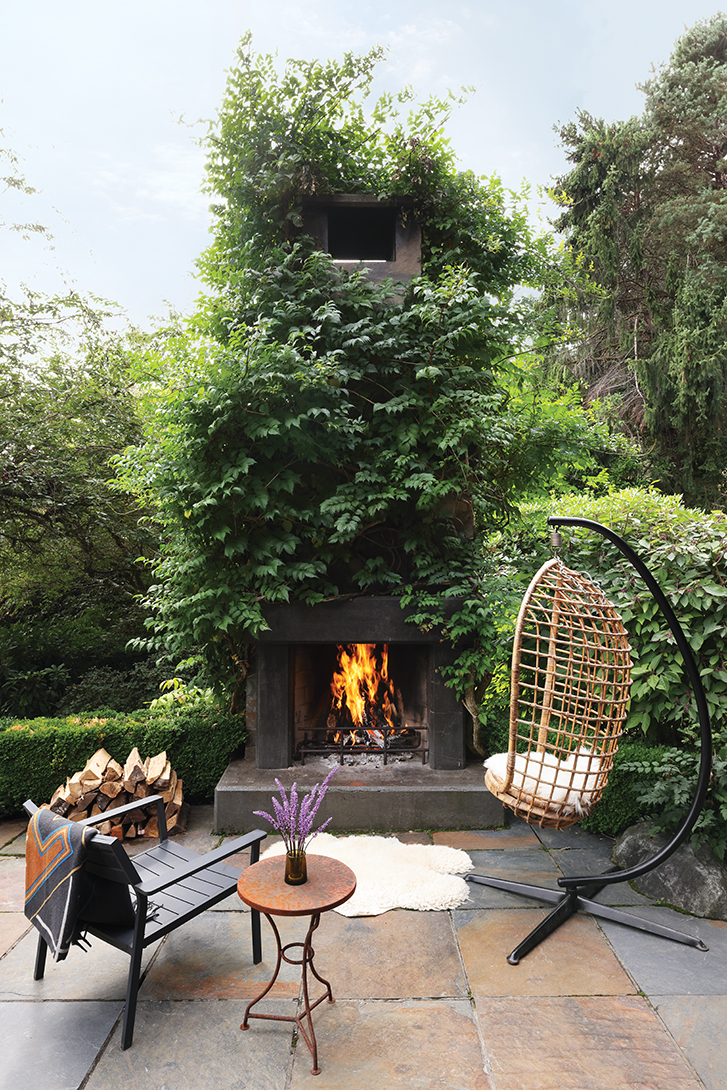 Outdoor patio with a tall fireplace covered in greenery, featuring a hanging chair, a lounge chair, and a small table.