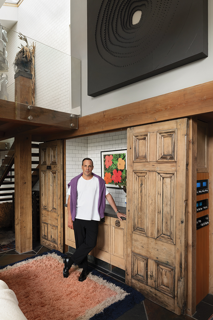 A man standing in a rustic modern interior with wooden beams, a large black artwork, and a colourful print on the counter.