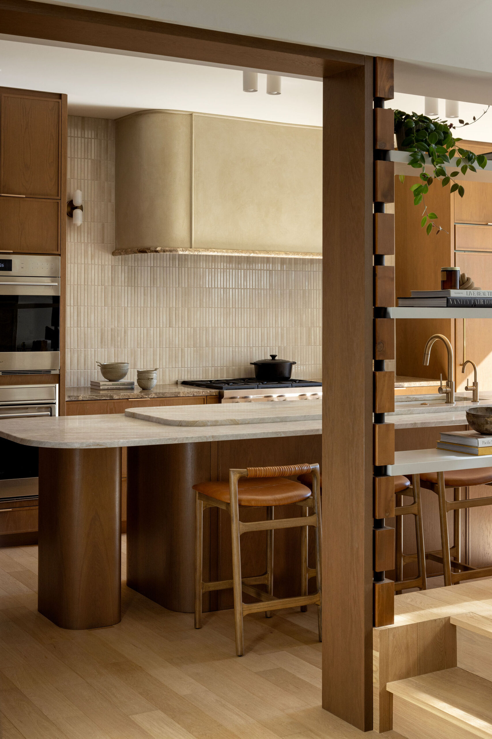 Kitchen with long stone island, oak cabinetry, curved plaster range hood and custom open shelving dividing the kitchen and living area