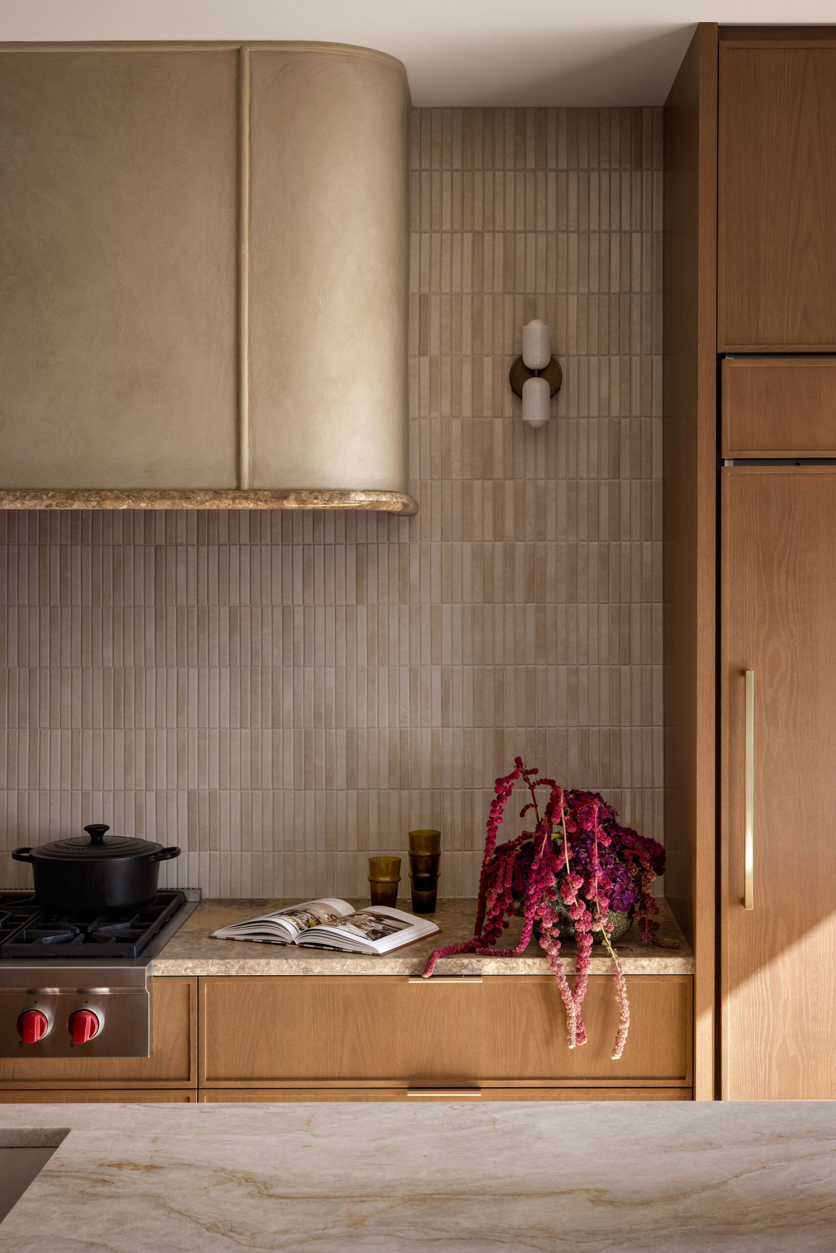 Close-up view of kitchen with long stone island, oak cabinetry, ribbed tile backsplash and curved plaster range hood