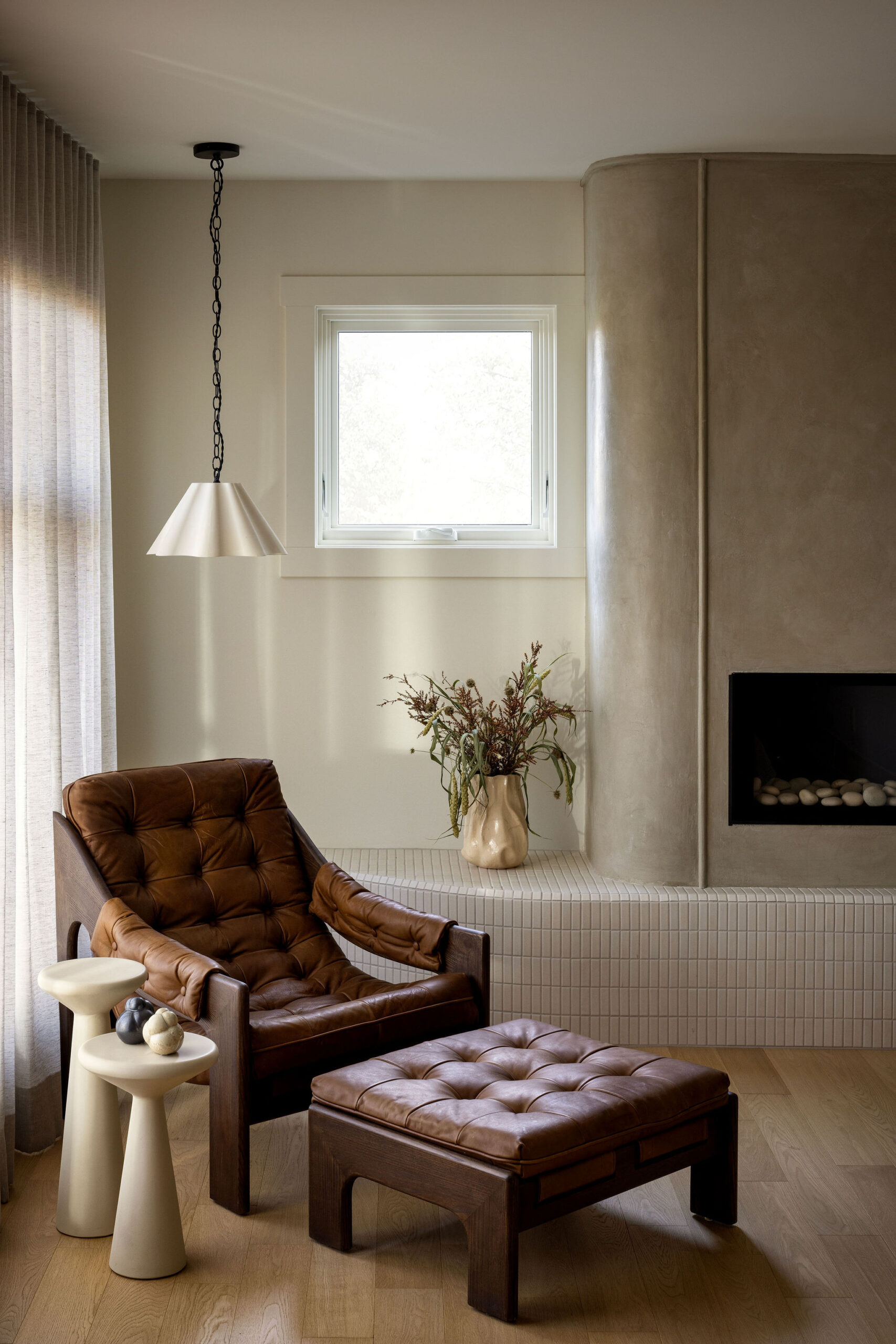 Bedroom sitting area with curved fireplace, brown leather lounge chair, hardwood flooring, pendant and natural light
