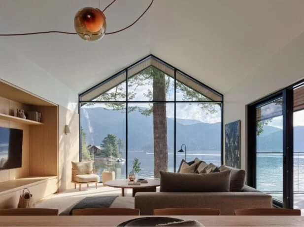 Interior view of living room with vaulted ceilings, floor-to-ceiling windows, neutral and wood furnishings, and ocean view.
