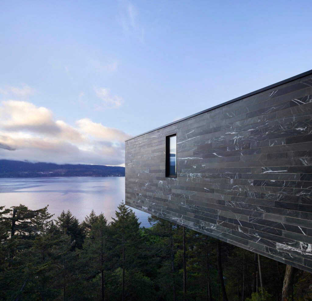 Side view highlighting the cantilevered upper level of the home overlooking trees and the water