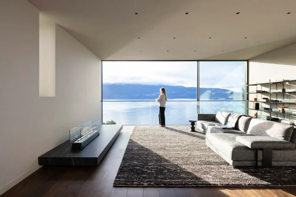 Woman in living room with panoramic views of the ocean, featuring a minimalist fireplace, light grey sectional, grey rug and full-height glazing with glass guardrail