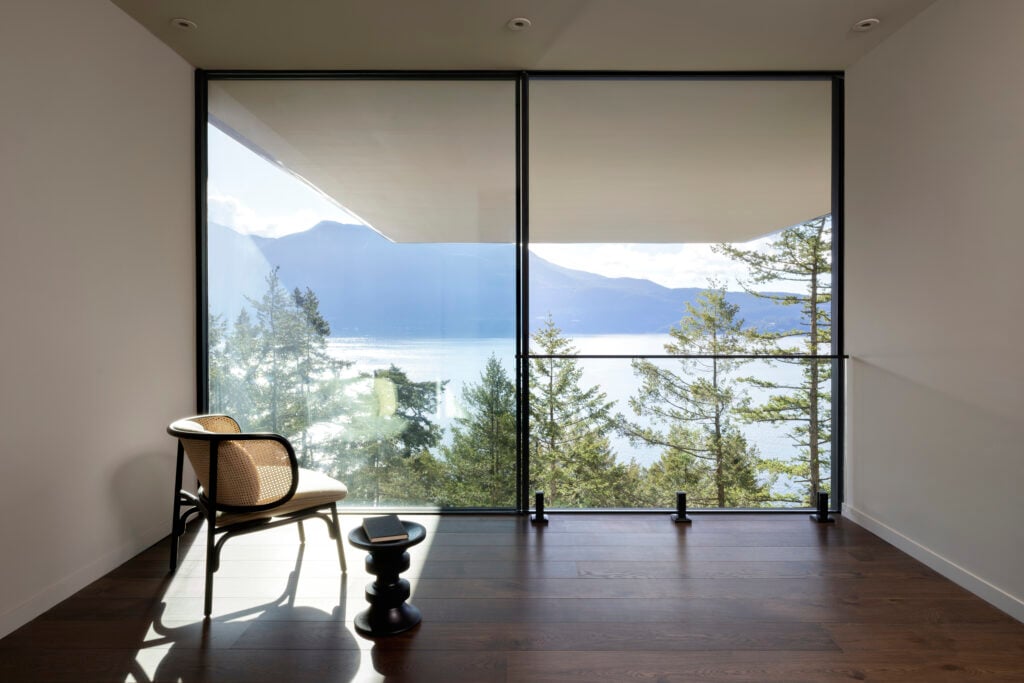 Lower level bedroom with woven armchair and sculptural side table, facing floor-to-ceiling windows with forest and water views