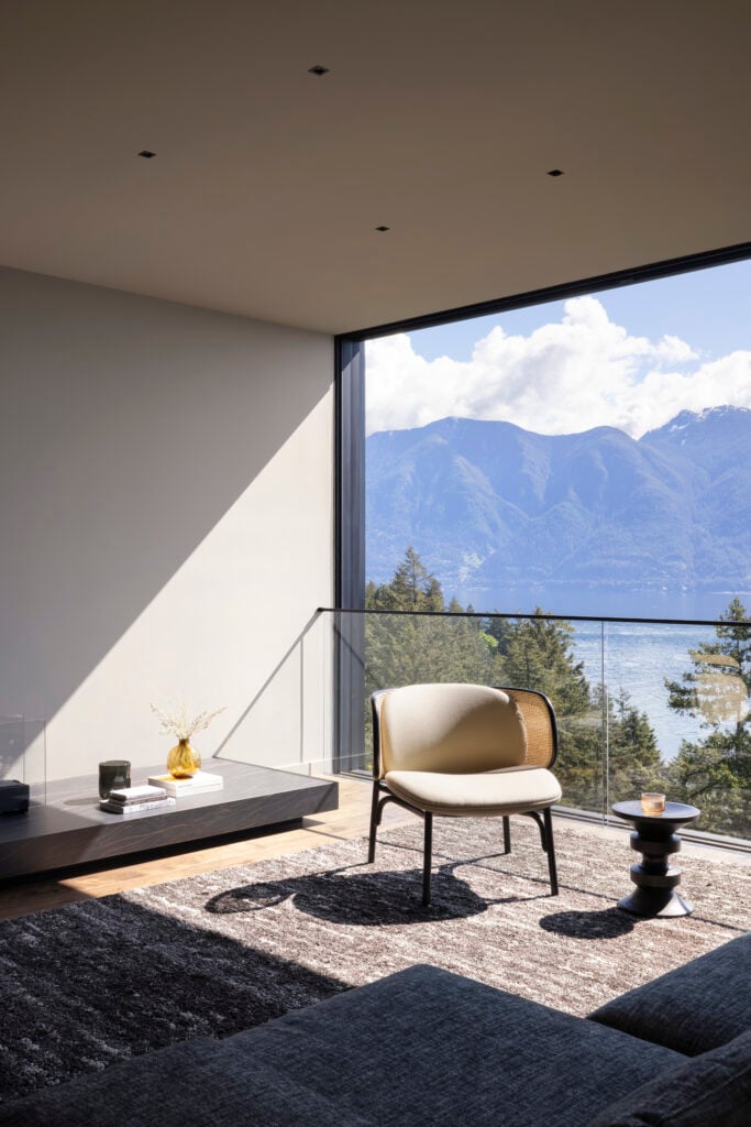 Close-up of living room corner with curved lounge chair and sculptural side table next to full-height window, set against ocean and mountain backdrop
