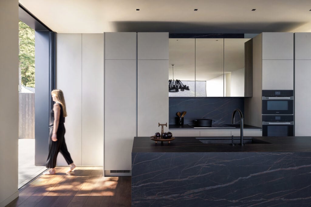 Minimalist kitchen with dark stone island, light grey cabinetry, and a woman walking out through a tall sliding glass door