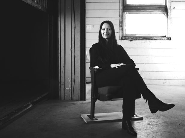 Black and white portrait of Tara-Lee Bennett sitting on a chair in an industrial space.